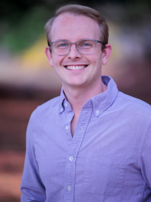 A medium close-up headshot photo of a light-skinned man wearing a purple button-up shirt and clear-rimmed glasses, with light-colored hair, smiling against a dark blurred background.