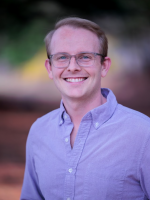 A medium close-up headshot photo of a light-skinned man wearing a purple button-up shirt and clear-rimmed glasses, with light-colored hair, smiling against a dark blurred background.