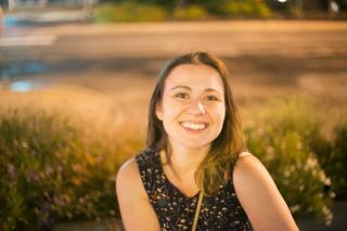 Headshot of Dr. Chicoli. She is wearing a sleeveless dress and smiling. Background is of bushes and greenery. 