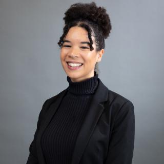 Image of Tara smiling at the camera. They are wearing a black turtleneck and black blazer, and they are framed within a gray background.